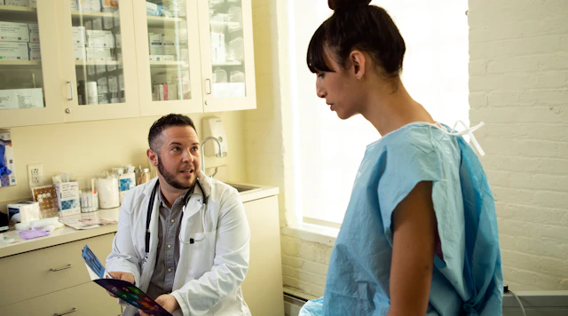 A transgender woman in a hospital gown speaking to her doctor a transgender man in an exam room
