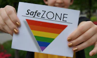 Image of person holding a sign featuring an upside-down triangle with rainbow bars. Text above triangle reads "Safe ZONE"