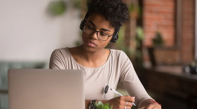 Young person wearing headphones and glasses on their laptop with a pencil.