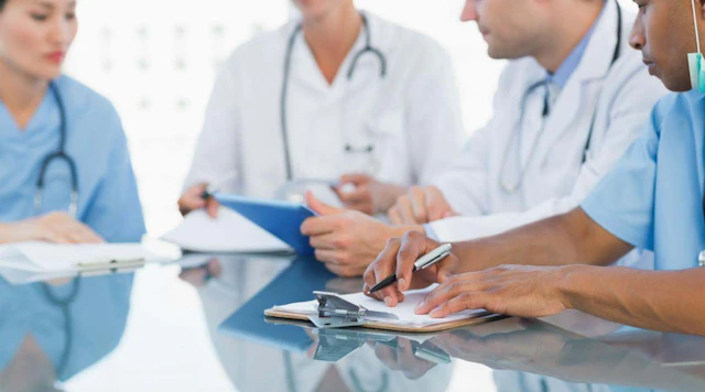 Four doctors and nurses surround a table with notepads and clipboards.