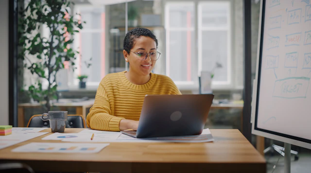 Black woman wearing glasses sitting at a table in an office working on a laptop