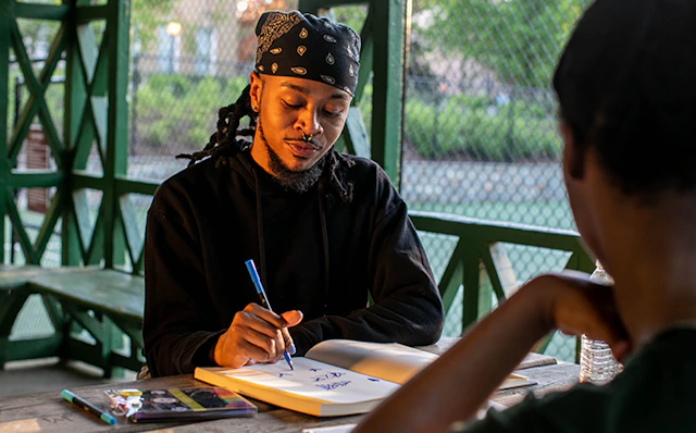 Two people sitting at a table. One person has their back to the camera. The other is looking down drawing in a sketchbook.